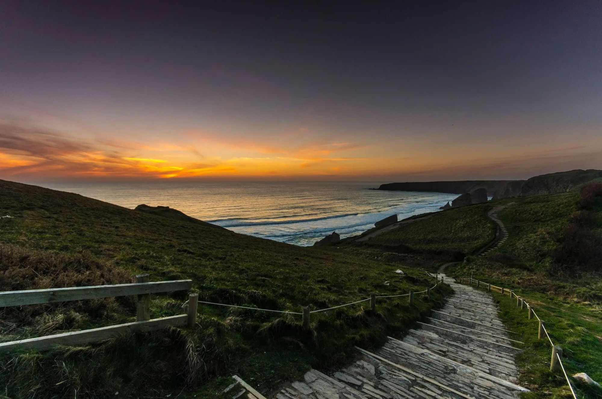 A fenced coast path running down the headland towards the sea at sunset